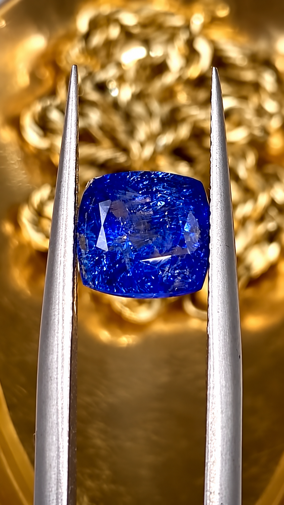 Deep blue sapphire gemstone held in jeweller's tweezers against a warm gold background